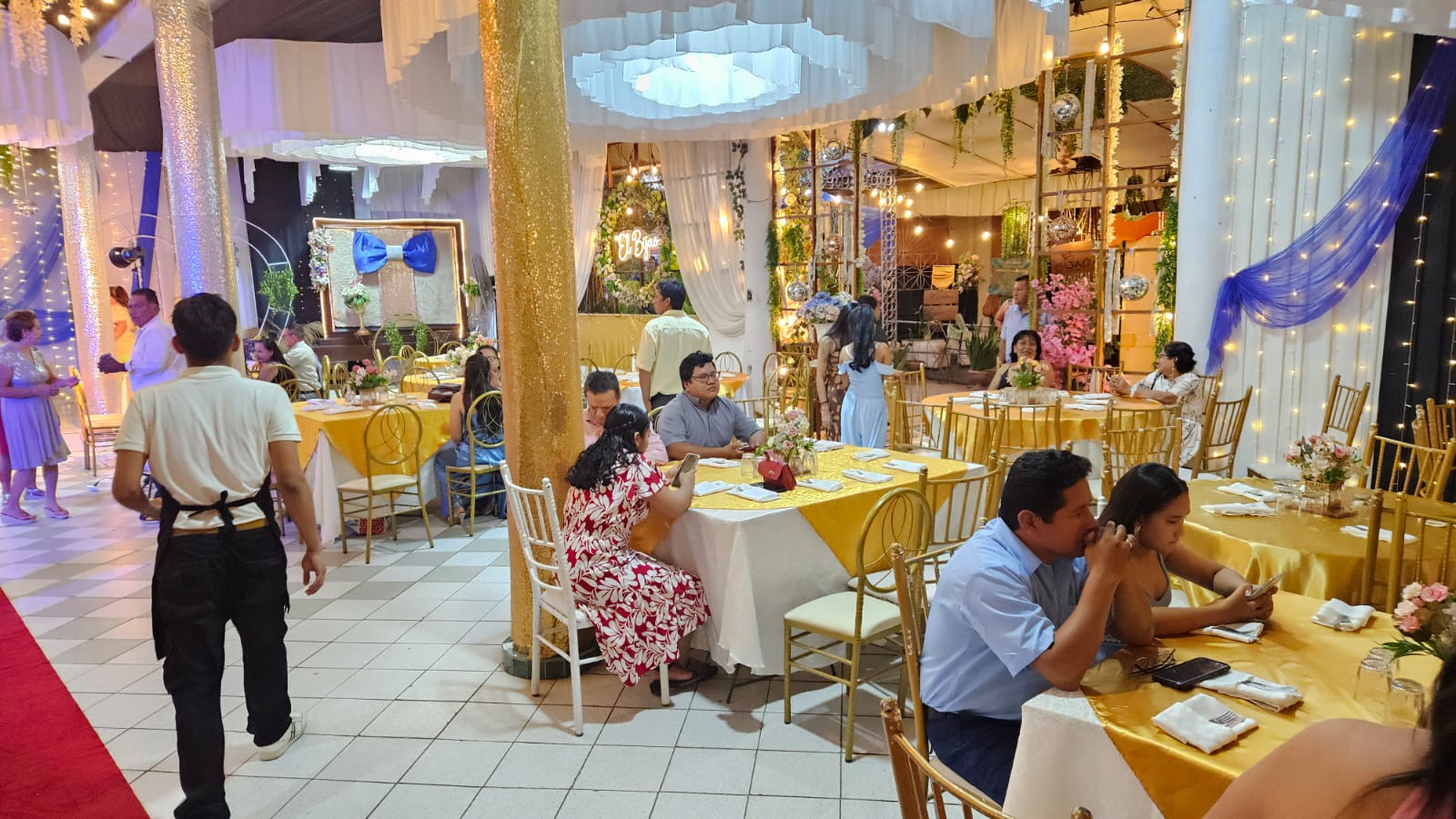 Salón montado con manteles dorados, cortinas blancas y luces cálidas durante una recepción