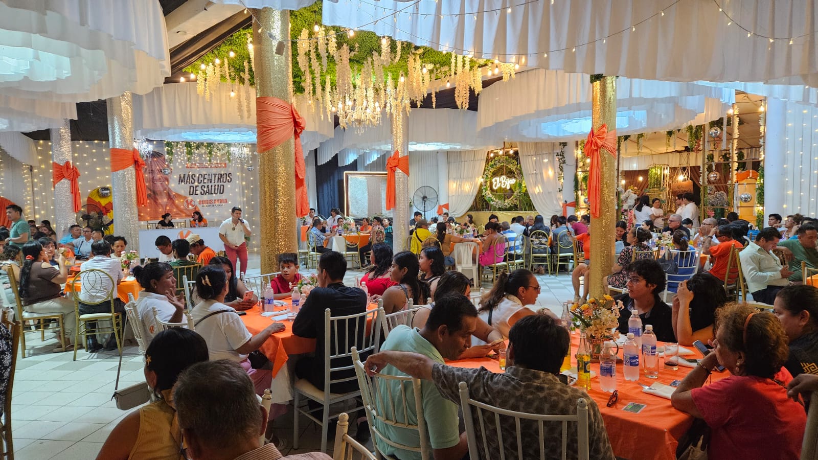 Evento masivo con mantelería naranja y decoración floral colgante en el salón