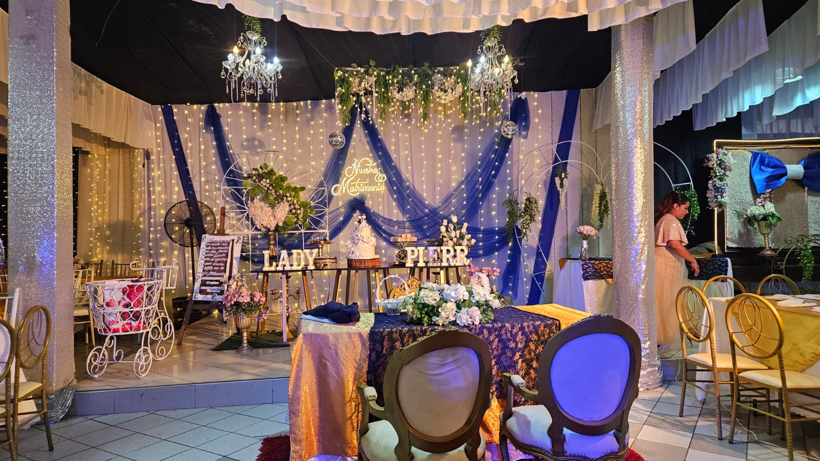 Mesa de novios con letrero Nuestro Matrimonio, cortinas azules y luces de hadas
